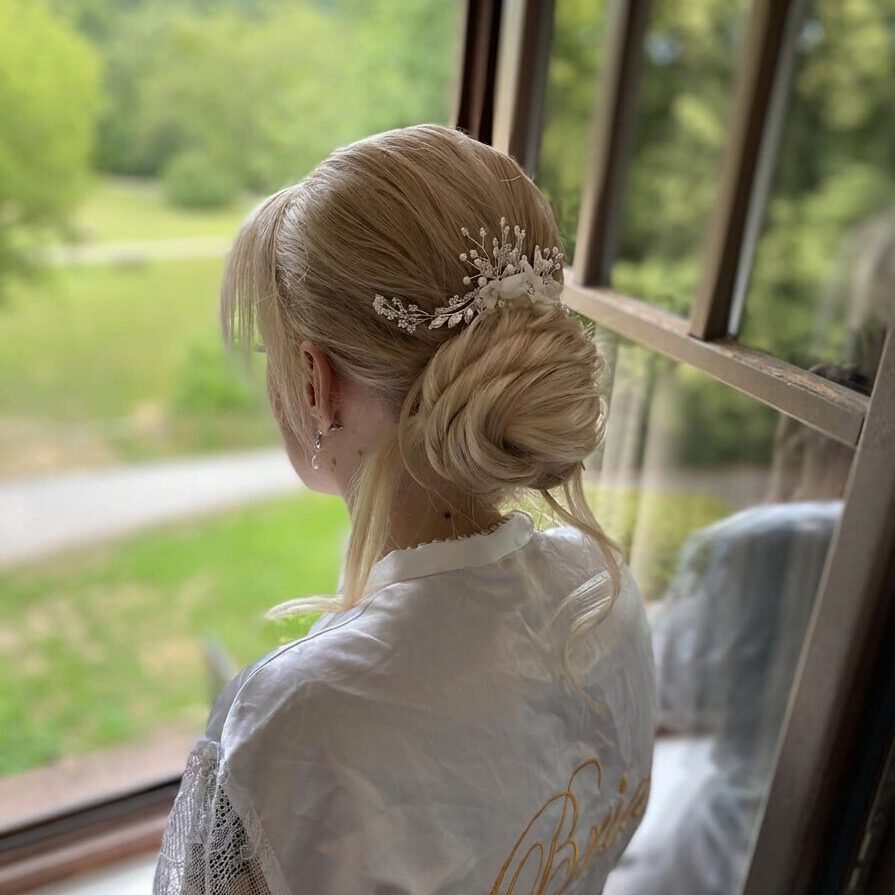 Braut mit blondem, elegantem Dutt und floralem Haarschmuck, romantisches Hochzeitsstyling am Fenster.